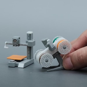 May include: Miniature grey 3D printer model with an orange platform and a second model with white, orange, and green components. The models are on a grey surface.