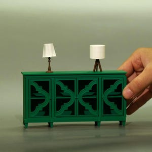 Può includere: Un mobile verde in miniatura con quattro ante e intagli decorativi, sormontato da due piccole lampade. Una lampada ha un paralume bianco e una base in bronzo, l'altra ha un paralume bianco e una base a treppiede in legno. Il mobile è tenuto in mano.