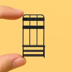 Könnte beinhalten: Ein Miniatur-Garderobenständer aus schwarzem Metall mit einer Bank und zwei Regalen. Der Garderobenständer hat drei Haken und eine geschwungene Oberseite. Das Stück wird zwischen zwei Fingern vor einem gelben Hintergrund gehalten.