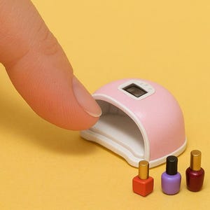 Può includere: Una lampada per unghie rosa con un display digitale del timer, aperta e pronta all'uso. Tre bottiglie in miniatura di smalto per unghie sono allineate davanti alla lampada: rosso, viola e granata.