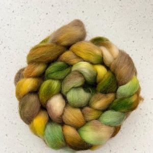 May include: A hand-spun yarn blend in shades of green, brown, and yellow. The yarn is arranged in a loose braid, creating a soft and textured look.