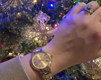 May include: A gold-toned wristwatch with a round face and Roman numeral hour markers. The watch has a linked metal band and is worn on a wrist. The background features a decorated Christmas tree with colorful lights and ornaments.