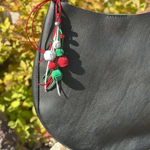 May include: Black leather handbag with a decorative tassel. The tassel features red, white, and green knotted accents, with a silver bead. The bag has a curved shape and a textured surface.