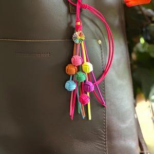 May include: A brown leather tote bag with a colorful tassel keychain. The keychain features a daisy charm and several knotted balls in various colors, including pink, orange, blue, and green. The bag has a pocket and a handle.