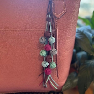 May include: A pink leather tote bag with a decorative tassel. The tassel features small, knotted balls in shades of blue, green, pink, and brown. The bag has a simple design with a single exterior pocket.