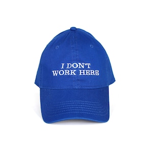 Peut inclure: Casquette de baseball bleu roi avec le texte blanc brodé "I DON'T WORK HERE". La casquette a une visière incurvée et un bouton sur le dessus. Le chapeau est de couleur unie et semble être fait d'un tissu durable.