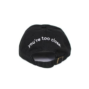 Könnte beinhalten: Schwarze Baseballkappe mit dem weißen Schriftzug "you're too close". Die Kappe hat einen gebogenen Schirm und einen verstellbaren Riemen mit Metallschnalle.