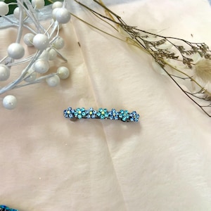 May include: A blue and turquoise floral hair clip. The clip features a row of small, sparkling flowers in varying shades of blue and turquoise. The flowers are set on a dark metal base, creating a delicate and elegant accessory.