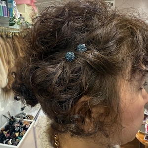 May include: Two small, round, blue crystal-encrusted hair pins are secured into a textured, curly brown hairstyle. The pins feature a sparkling, multi-faceted surface that catches the light, providing a decorative accent to the dark hair.
