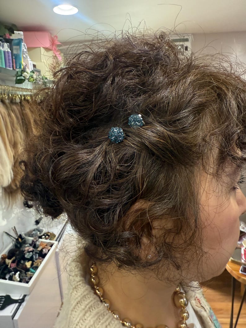 May include: Two small, round, blue crystal-encrusted hair pins are secured into a textured, curly brown hairstyle. The pins feature a sparkling, multi-faceted surface that catches the light, providing a decorative accent to the dark hair.