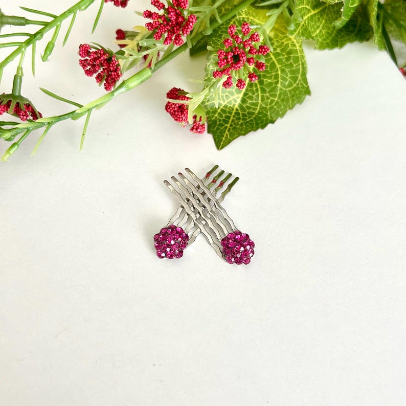 May include: Two silver-toned hair combs arranged in an X-shape, each featuring a spherical topper encrusted with small, vibrant fuchsia-pink crystals. The metal combs have a wavy design, and the fuchsia accents provide a bright, reflective finish.