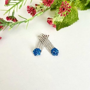 May include: Two silver-toned metal hair combs, each featuring a spherical cluster of small, vibrant blue rhinestones at the top. The combs have wavy teeth and are positioned side-by-side, showcasing the metallic finish and the blue crystal accents.