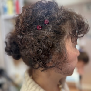 May include: Two small, round hair pins featuring clusters of deep red, faceted crystals. The pins are secured into dark brown, curly hair, with the vibrant red stones providing a distinct contrast against the dark texture of the hair strands.