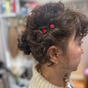 May include: Two small, round, red crystal-encrusted hair pins are secured into dark, curly, textured hair. The pins feature a cluster of tiny, sparkling red stones arranged in a spherical shape, providing a vibrant contrast against the dark hair.