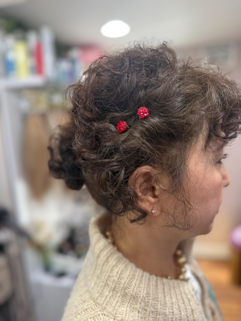 May include: Two small, round, red crystal-encrusted hair pins are secured into dark, curly, textured hair. The pins feature a cluster of tiny, sparkling red stones arranged in a spherical shape, providing a vibrant contrast against the dark hair.