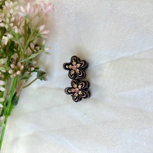 May include: A pair of flower-shaped earrings with a dark bronze finish. Each earring has a central pink gemstone surrounded by smaller clear gemstones. The earrings are on a white, textured background, with pink flowers in the top left corner.