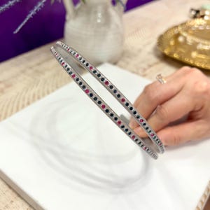 Op de afbeelding: Twee zilveren hoofdbanden met zwarte en roze strass steentjes. De hoofdbanden zijn dun en hebben een eenvoudig ontwerp.