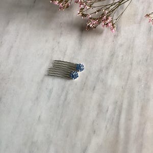 May include: A silver-toned hair comb featuring two round, blue crystal-encrusted spheres attached to the top. The comb has multiple wavy metal teeth designed for securing hair, with the blue spheres providing a decorative, textured finish.