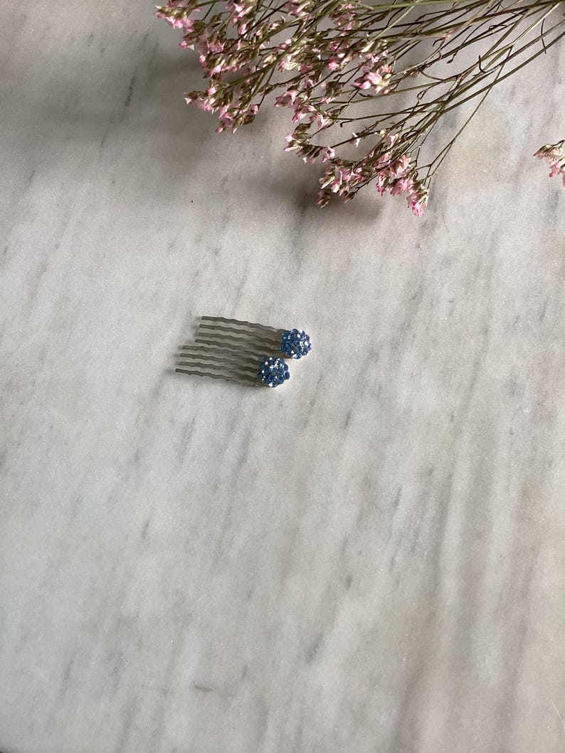 May include: A silver-toned hair comb featuring two round, blue crystal-encrusted spheres attached to the top. The comb has multiple wavy metal teeth designed for securing hair, with the blue spheres providing a decorative, textured finish.