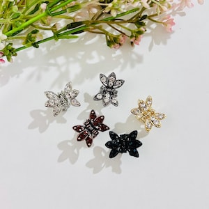 May include: Five small, flower-shaped hair clips in silver, black, gold, and burgundy. Each clip features a rhinestone center and petals. The clips are arranged on a white surface with greenery and pink flowers in the background.