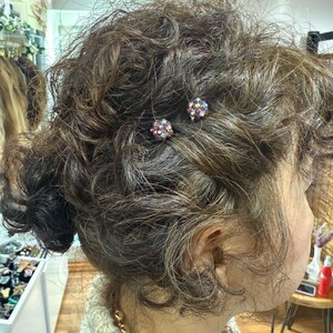 May include: Two small, round hair pins featuring a cluster of dark purple and iridescent multicolored crystals. The pins are secured into dark brown, curly hair, with the spherical crystal embellishments visible against the textured hair strands.