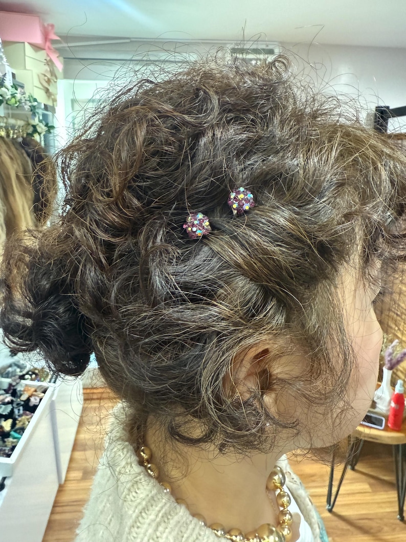 May include: Two small, round hair pins featuring a cluster of dark purple and iridescent multicolored crystals. The pins are secured into dark brown, curly hair, with the spherical crystal embellishments visible against the textured hair strands.