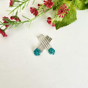 May include: Two silver-toned hair combs featuring wavy metal teeth and a circular cluster of bright turquoise rhinestones at the top of each piece. The combs are positioned in an overlapping cross shape against a plain white background.