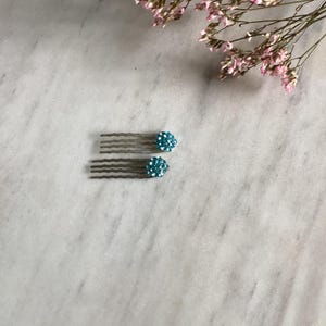 May include: Two hair combs featuring a metallic silver-toned base with five wavy teeth. Each comb is topped with a round, dome-shaped embellishment covered in small, sparkling light blue crystals that catch the light, creating a textured appearance.