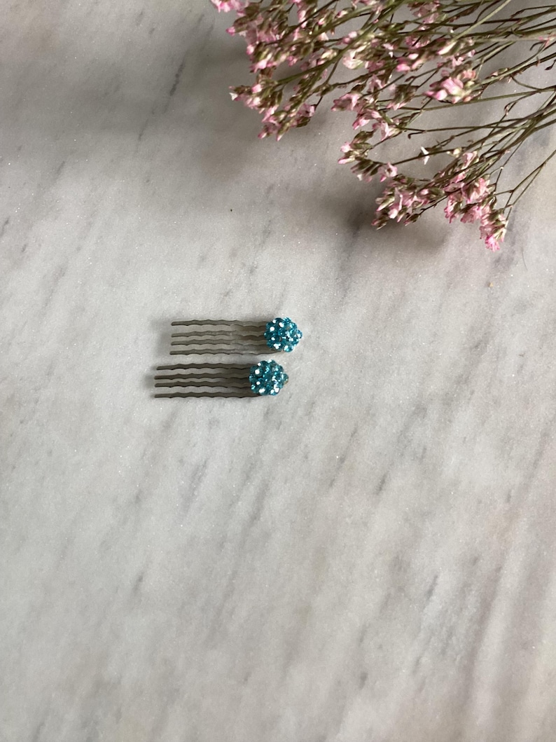 May include: Two hair combs featuring a metallic silver-toned base with five wavy teeth. Each comb is topped with a round, dome-shaped embellishment covered in small, sparkling light blue crystals that catch the light, creating a textured appearance.