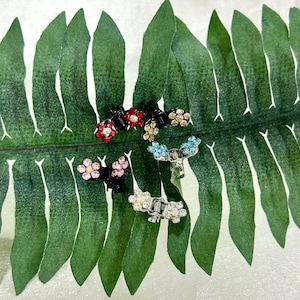 Pode incluir: Uma coleção de pequenas presilhas de cabelo em forma de flor em várias cores, incluindo vermelho, rosa, azul e transparente, com detalhes em strass. As presilhas são pretas e prateadas. As presilhas estão dispostas em uma grande folha verde.
