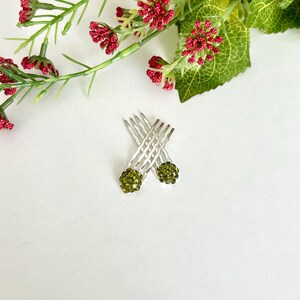 May include: Two silver-toned hair combs featuring a cluster of small, olive-green rhinestones at the top of each piece. The combs are arranged in a crossed position, showcasing the metallic teeth and the sparkling, circular stone embellishments.