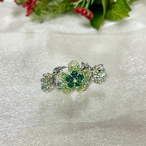 May include: A silver-toned hair clip featuring a floral design. The flower is adorned with green and yellow-green rhinestones, with smaller, iridescent stones on the side accents. The clip is set against a white, textured background.
