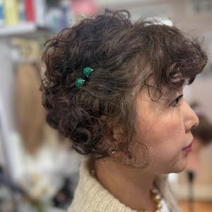 May include: Two small, round, textured green hair pins are secured in short, dark brown, curly hair. The pins feature a vibrant emerald green surface with a subtle, bumpy texture, providing a distinct contrast against the dark, wavy hair strands.