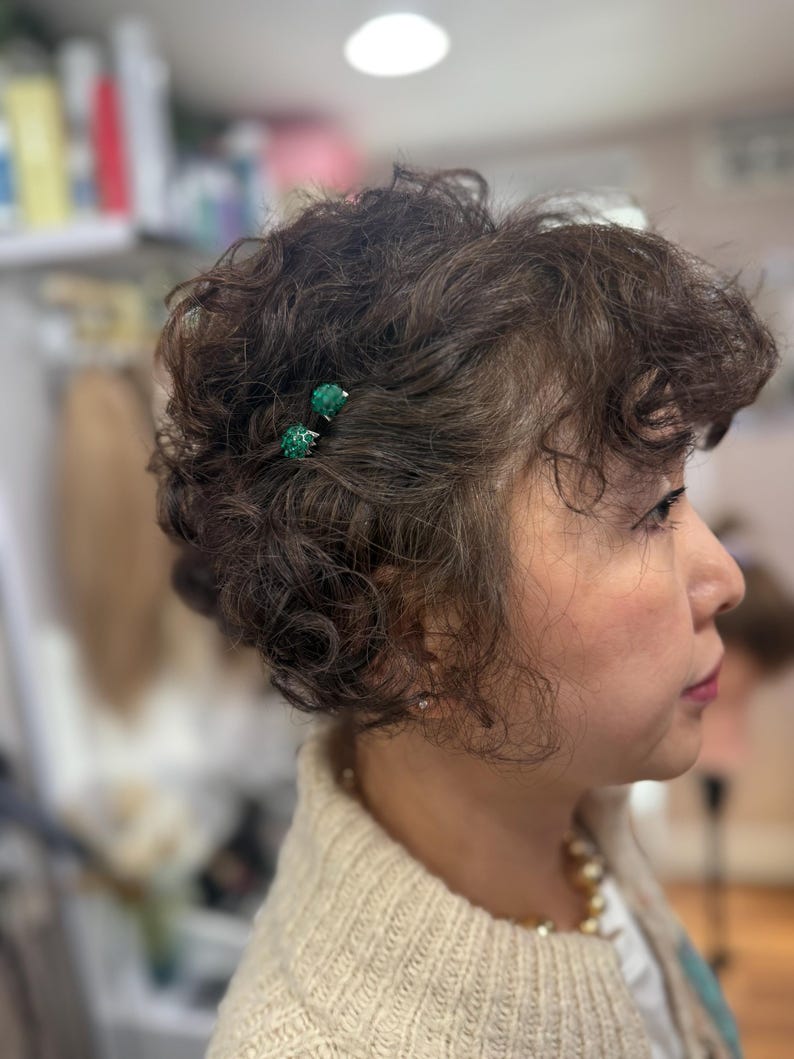 May include: Two small, round, textured green hair pins are secured in short, dark brown, curly hair. The pins feature a vibrant emerald green surface with a subtle, bumpy texture, providing a distinct contrast against the dark, wavy hair strands.