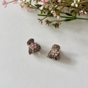 Puede incluir: Dos pequeñas pinzas para el cabello de metal en tono bronce antiguo con un intrincado patrón de filigrana. Cada pinza está adornada con un cristal iridiscente central rodeado de pedrería redonda transparente en forma de flor.