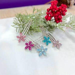 May include: Four silver-toned hair combs, each featuring a five-petaled flower ornament encrusted with small sparkling rhinestones in shades of light pink, dark pink, bright blue, and light blue, arranged on a white surface with greenery.