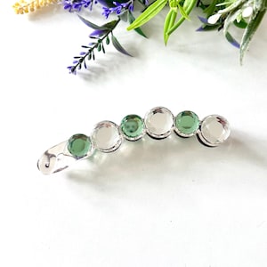 Peut inclure: Une barrette à cheveux incurvée ornée d'une série de six pierres circulaires à facettes disposées en ligne. Les pierres alternent entre du verre transparent incolore et du verre vert sauge translucide, montées sur une monture en métal argenté.