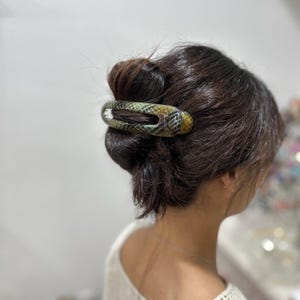 Op de afbeelding: Een langwerpige, ovaalvormige haarclip met een gestructureerd slangenleerpatroon in tinten groen, bruin en zwart. Het accessoire is voorzien van een centrale uitsparing en is horizontaal bevestigd over een donkerbruine haarknot voor stevigheid.