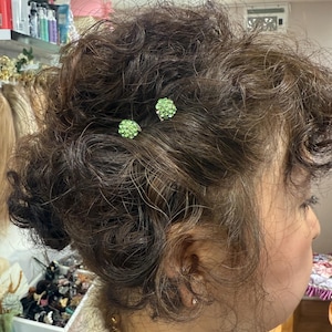 May include: Two small, round hair pins featuring clusters of light green rhinestones are secured into dark brown, curly hair. The pins have a metallic base and are positioned near the crown of the head, adding a subtle sparkle to the hairstyle.