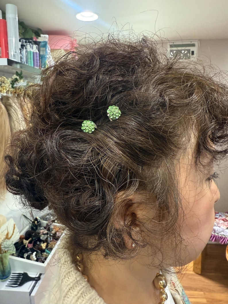 May include: Two small, round hair pins featuring clusters of light green rhinestones are secured into dark brown, curly hair. The pins have a metallic base and are positioned near the crown of the head, adding a subtle sparkle to the hairstyle.