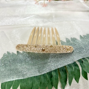 May include: A decorative hair comb with a beige base and a row of seven long, pointed teeth. The comb's base is embellished with small, sparkling rhinestones in shades of gold and clear, creating a glamorous accessory.