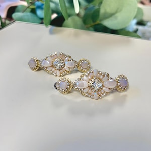 May include: Two ornate hair clips with a gold-tone setting. Each clip features a central square-cut clear stone surrounded by a floral design of white and pink stones. The clips have a barrette style clasp.