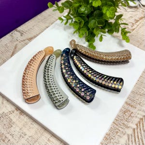 May include: Five curved banana hair clips arranged in a row. Each clip features a distinct colour base including peach, light grey, dark blue, and brown, and is embellished with rows of sparkling, multi-faceted rhinestones along the outer surface.