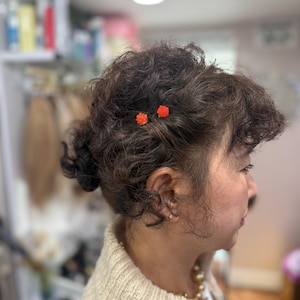 May include: A side profile view of dark brown curly hair styled in an updo, accented with two small, bright orange flower-shaped hair pins. The hair is textured with soft curls, and the pins are positioned near the temple area of the head.