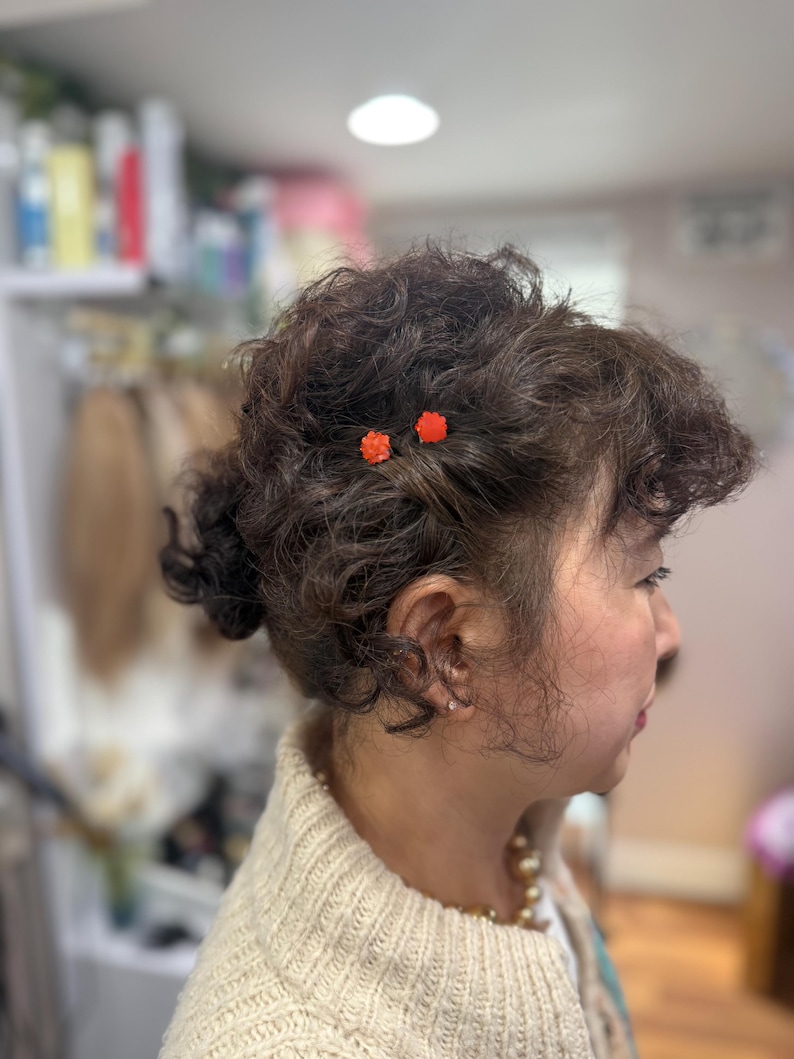 May include: A side profile view of dark brown curly hair styled in an updo, accented with two small, bright orange flower-shaped hair pins. The hair is textured with soft curls, and the pins are positioned near the temple area of the head.