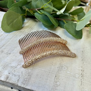May include: Two rose gold hair combs featuring a curved top edge encrusted with small, shimmering clear crystals. The metal teeth are thin and wavy, designed to grip hair securely. The finish on the metal is a metallic rose gold tone.