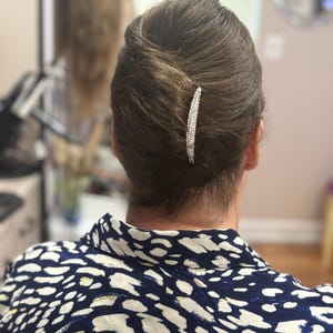 Op de afbeelding: Een kapsel in een lage knot, vastgezet met een sprankelende, zilveren haarspeld. De haarspeld is versierd met talrijke kleine, heldere kristallen. De vrouw draagt een top met een marineblauw en wit patroon.