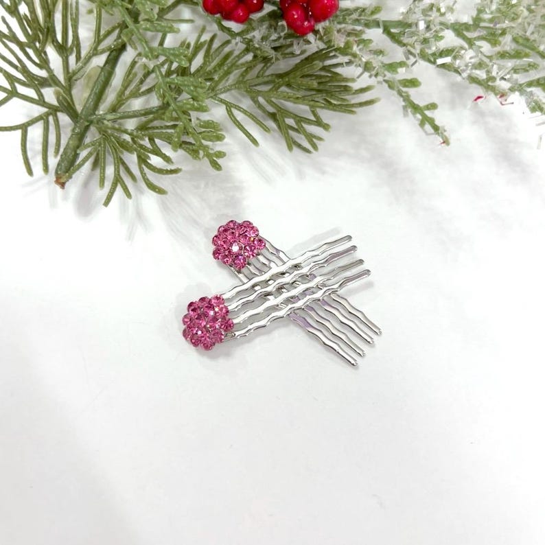 May include: A pair of silver-toned hair combs featuring rounded ends encrusted with small, vibrant pink crystals. The combs have wavy, multi-pronged teeth designed for securing hair, with the pink crystal embellishments positioned at the top.