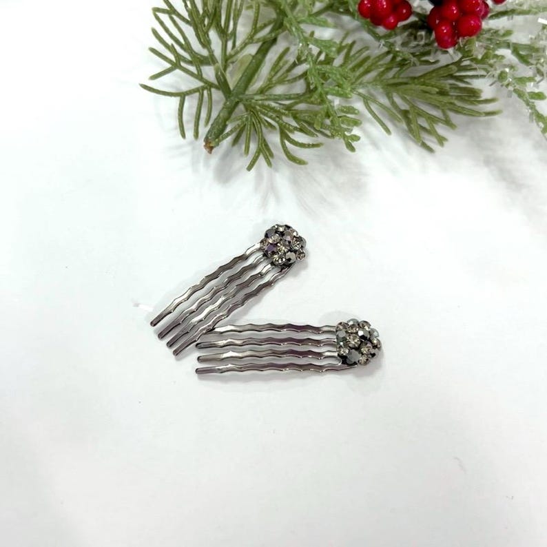 May include: Two silver-toned hair combs with wavy teeth, each featuring a decorative cluster of small, sparkling clear crystals at the top. The combs are arranged in a V-shape, showcasing the metallic finish and the faceted crystal embellishments.