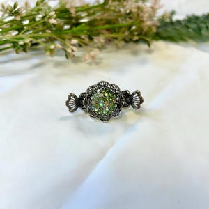 Könnte beinhalten: Ein silberfarbener Haarklammer mit einem zentralen Blumendesign, das mit grünen Strasssteinen besetzt ist. Die Klammer hat dekorative Seitenakzente und ist vor einem weißen Hintergrund mit Grün angeordnet.